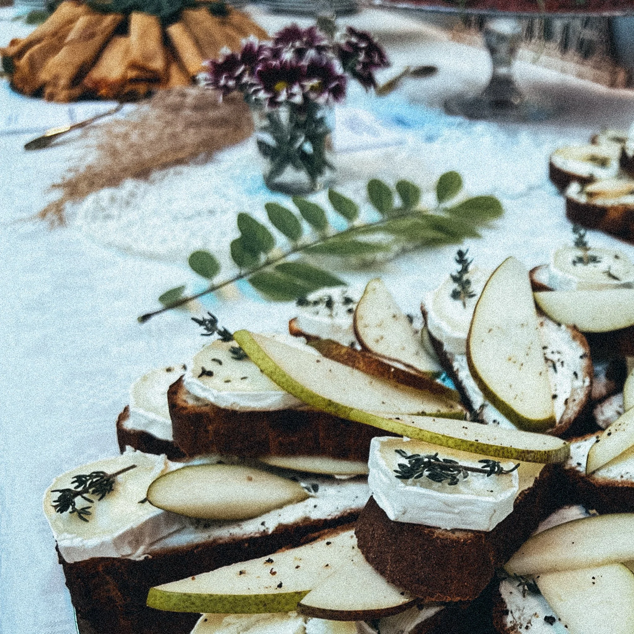 Tisch, belegt mit Baguette-Scheiben, die mit Brie und Birnenscheiben garniert sind. Im Hintergrund ein gestreifter Strauß aus lila-weißen Blumen und ein Teller mit Gebäck.