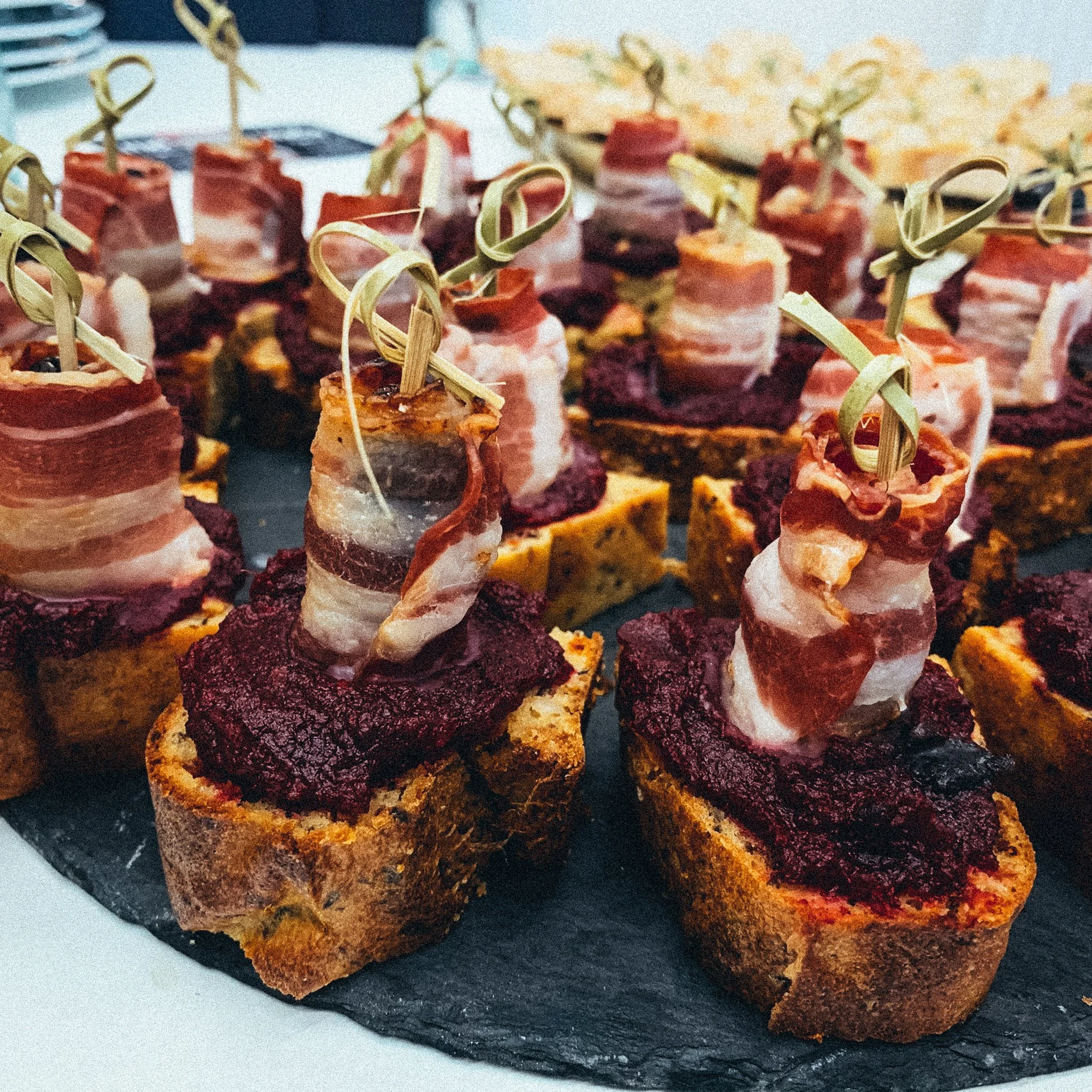 Canapés mit Brot, Roter Beete-Aufstrich und umwickeltem Speck, dekorativ angerichtet auf einem Tablett.