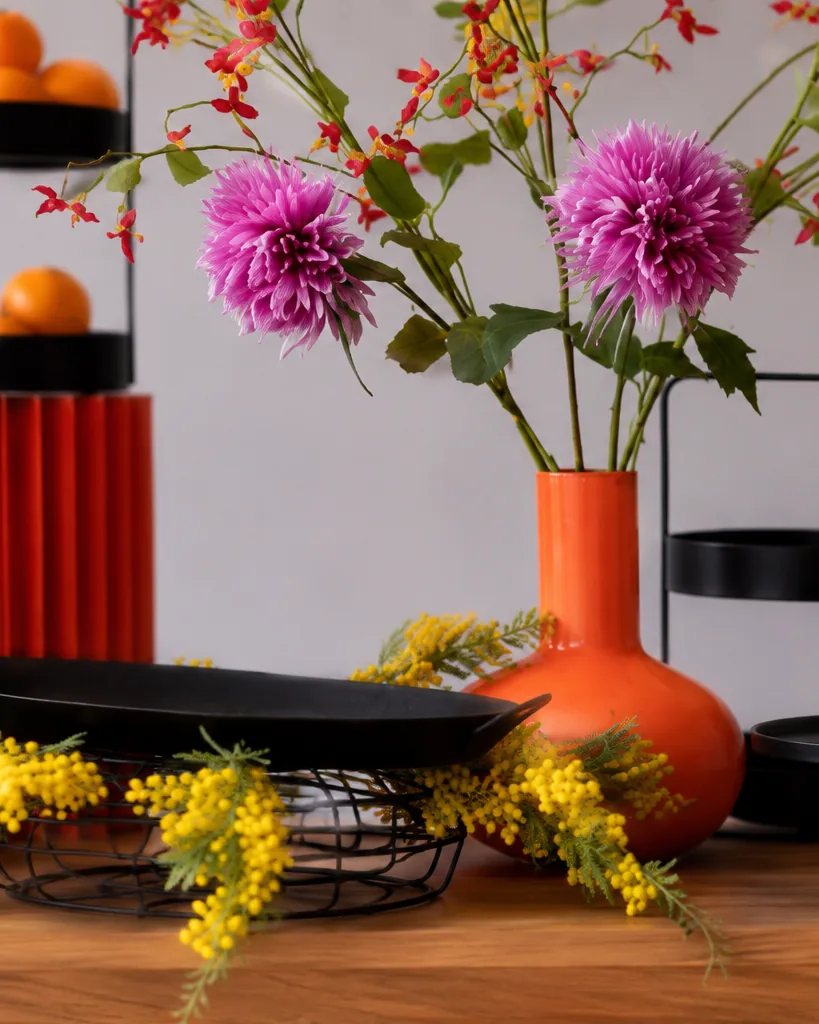 Nahaufnahme: rote Riffelvase, orangefarbene Vase und schwarze Schalen, dazu Dahlien, Mimose und Zitrusfrüchte.