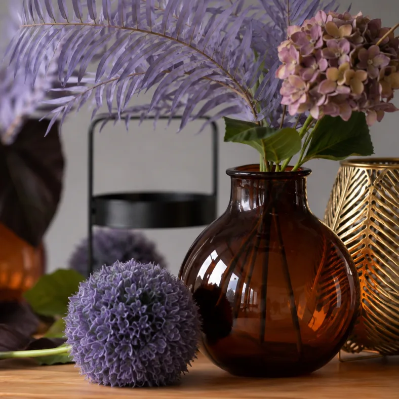 Nahaufnahme einer Alliumkugel neben der Bauchvase aus Bernsteinglas mit altrosa Hortensie, dahinter eine goldene Blattvase.
