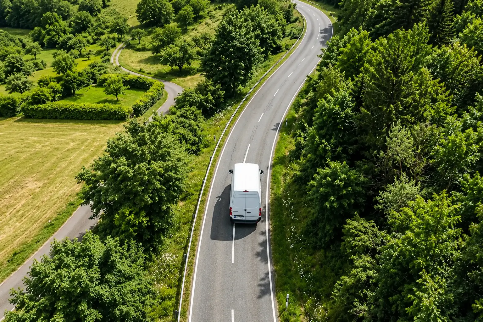 Weißer Transporter von oben auf einer geschwungenen Landstraße zwischen Wiesen und Bäumen