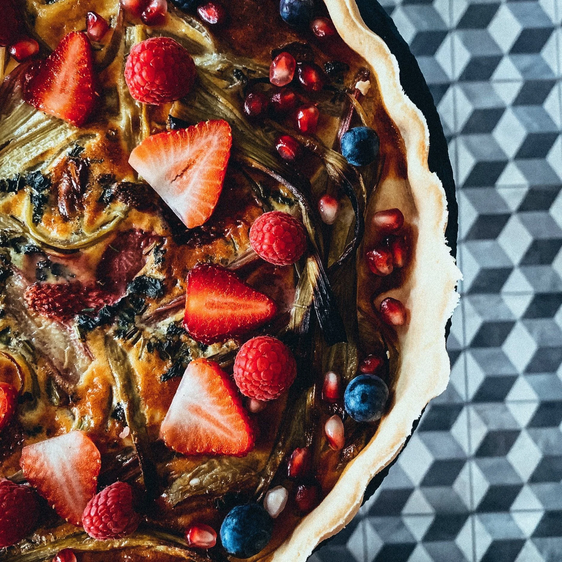 Obsttarte mit Erdbeeren, Himbeeren, Heidelbeeren und Granatapfelkernen auf geometrisch gemustertem Hintergrund.