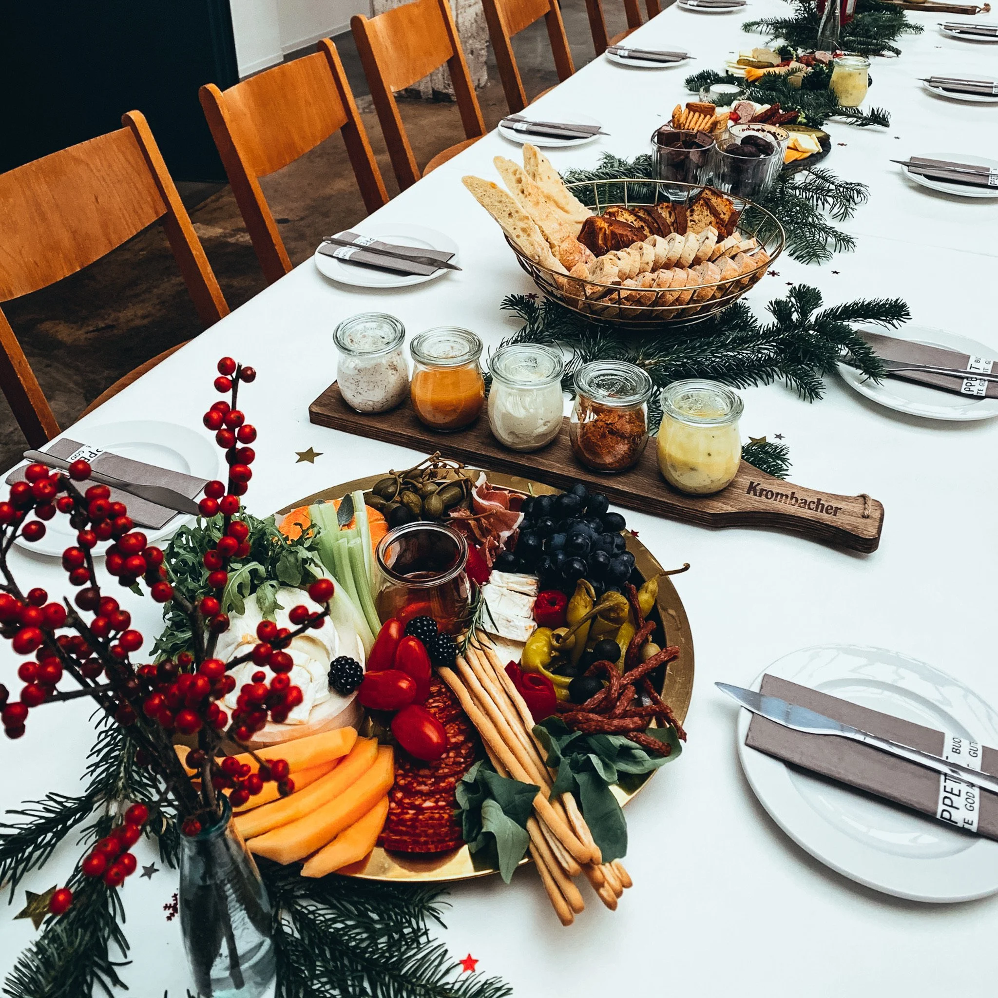 Festlich gedeckter Tisch mit rustikaler Dekoration. Es gibt Teller mit Brotauswahl, Gläser mit Dips und diverse Vorspeisen wie Käse, Früchte und Gemüse. Der Tisch ist mit Tannenzweigen und roten Beeren geschmückt.